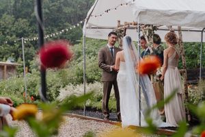 Outdoor wedding ceremony under a white tent with string lights, bride in veil, groom in brown suit, and bridesmaids.