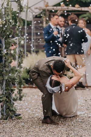 Groom dips bride for a romantic kiss as white confetti falls during outdoor wedding ceremony celebration.
