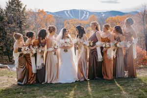 Bride with bridesmaids in earth-tone gowns holding bouquets with autumn mountain backdrop.