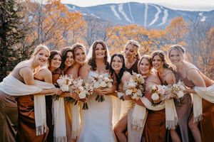 Bride poses with bridesmaids in rust and champagne gowns holding white bouquets with mountain backdrop.