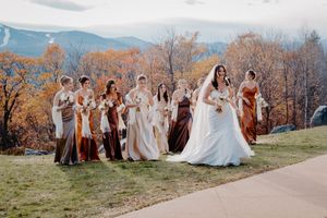 Bridesmaids in rust and bronze gowns walk with bride in white dress against fall mountain backdrop.