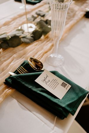 Elegant wedding place setting with green napkin, gold cutlery, menu card with QR code, and crystal champagne flute.