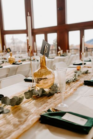 Elegant wedding reception table with amber vase centerpiece, green napkins, eucalyptus runner, and table number 6.