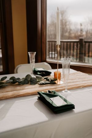 Elegant wedding table setting with green napkins, gold candlestick, eucalyptus runner, and crystal champagne flutes on white linen.