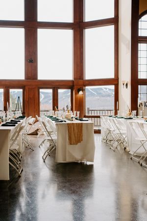 Elegant wedding reception hall with white tablecloths, green accents, and floor-to-ceiling windows overlooking snowy mountains.
