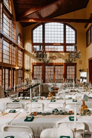 Grand wedding reception venue featuring vaulted ceilings, ornate chandeliers, large windows, and long tables with green napkins.