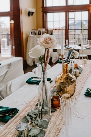 Elegant wedding reception table with white roses, greenery, amber bottles, and emerald green napkins on a white linen runner.