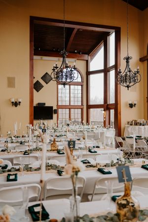 Wedding reception hall with long white tables, green napkins, chandeliers, and tall windows with natural light.