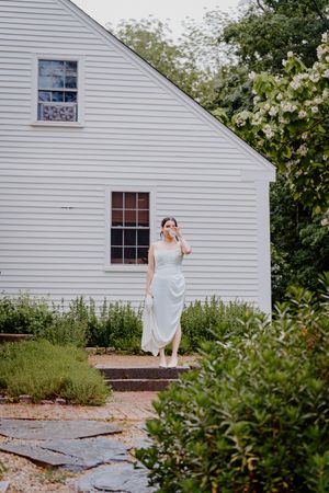 Bride in a white satin gown walks down stone steps outside a charming white New England farmhouse.