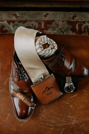 Groom wedding day details featuring brown leather dress shoes, flask engraved Sam, cigar, tie, and watch.