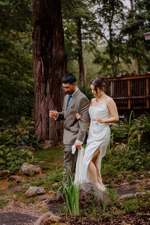 Bride and groom walking through lush garden path, groom in tan suit holding drink, bride in elegant white gown.