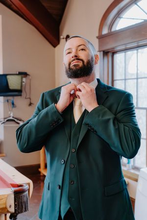 Bearded groom in emerald green suit and vest adjusting his gold tie while getting ready for his wedding.