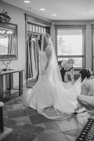 Black and white photo of bride in wedding gown with veil as attendants adjust her dress train.