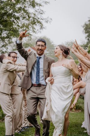 Joyful newlyweds celebrate with wedding party in a lush garden, groom raising hand in excitement.
