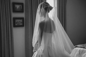 Black and white photo of a bride in a wedding dress and veil standing by a window on her wedding day.