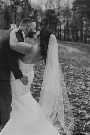 Black and white romantic photo of bride and groom sharing a kiss outdoors, with her pearl-dotted veil flowing behind.