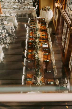 Aerial view of elegant wedding reception with long wooden farm table adorned with greenery garland and white chair settings.