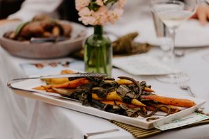 Elegant table setting with roasted carrots and vegetables on a white platter, wine glass, and pink flowers.