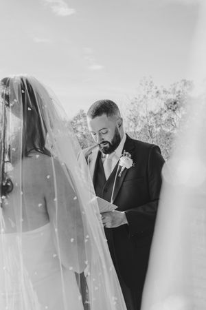 Black and white photo of bearded groom reading vows beside veiled bride at outdoor wedding ceremony.