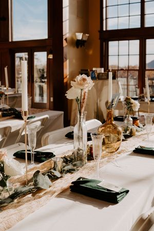 Elegant fall wedding reception table with green napkins, white flowers, gold bottles, and tall taper candles.