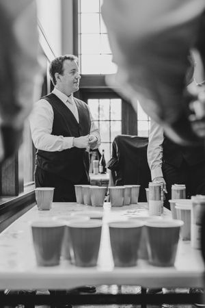 Black and white photo of a bartender preparing drinks with rows of cups on a bar counter.