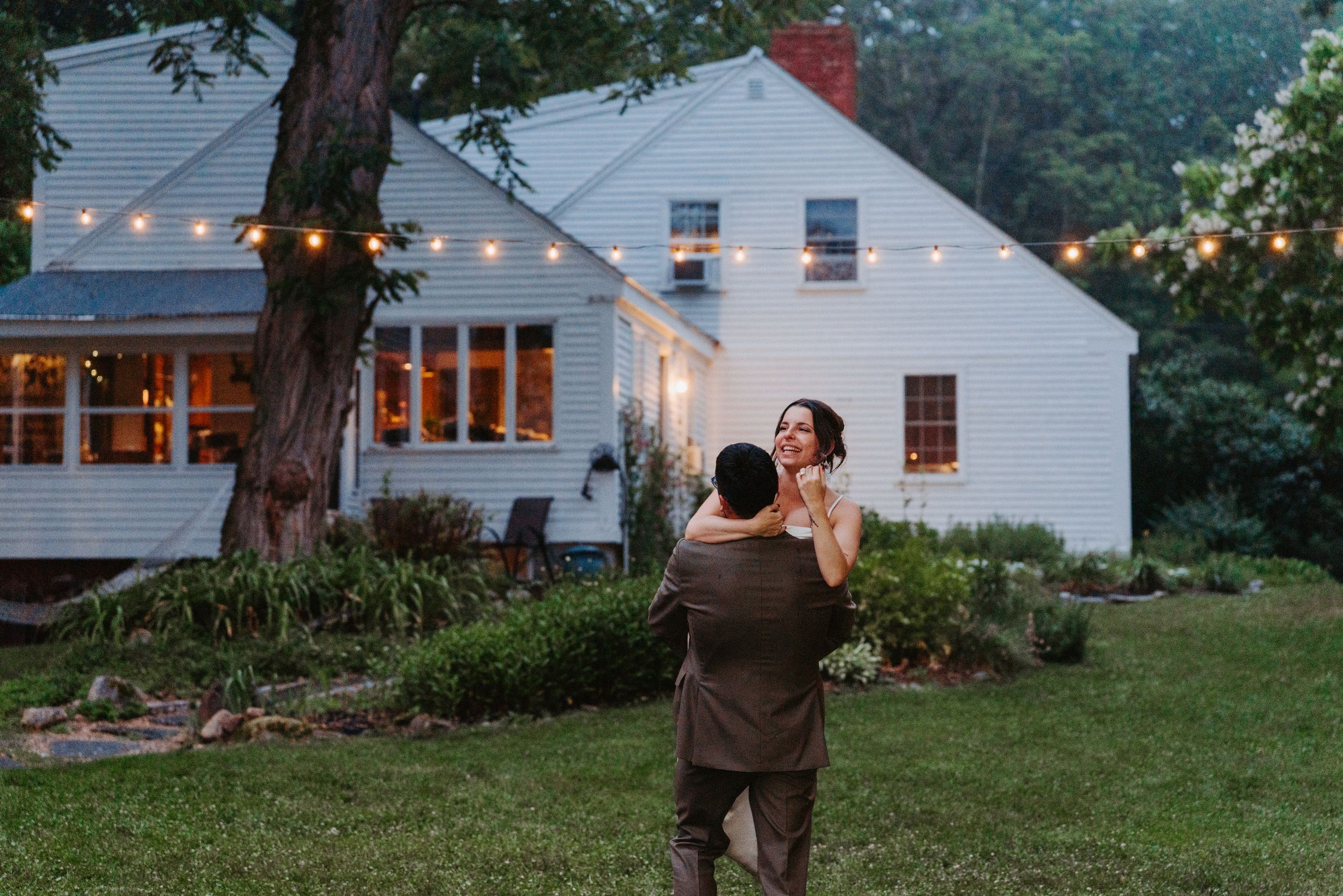 Couple dancing in a lush backyard at dusk with string lights and a charming white farmhouse in the background.