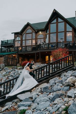Bride and groom walk rocky steps before a grand lodge venue with large windows and autumn foliage at dusk.