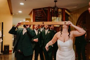 Bride and groomsmen in green suits doing the limbo with a wooden board at wedding reception.