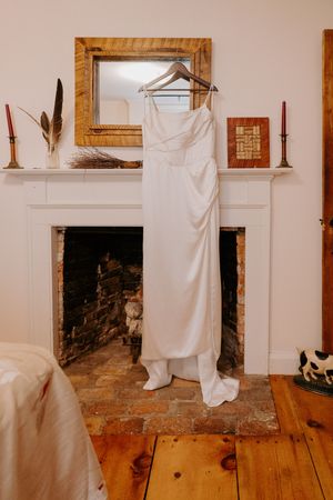 Elegant white satin wedding dress hanging on a hanger over a rustic fireplace mantel in a cozy room.