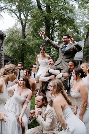 Joyful wedding party group photo with bride and groom elevated above bridesmaids in white dresses and groomsmen outdoors.
