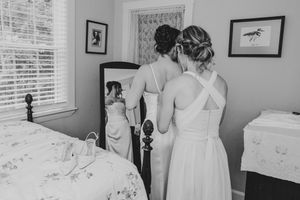 Black and white photo of two women in elegant dresses, one helping the other prepare in a cozy bedroom with a mirror.