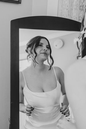 Black and white portrait of a bride in a white dress admiring herself in a full-length mirror.