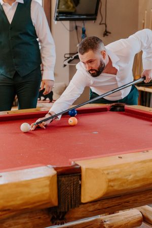 Bearded man in white shirt leaning over red pool table taking a shot with billiard cue stick.