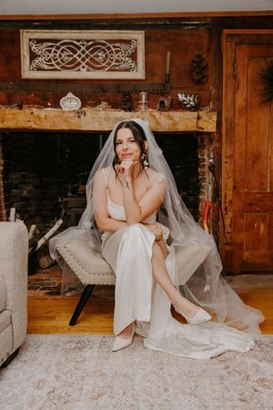 Bride in white gown and veil sits gracefully in chair before rustic stone fireplace.
