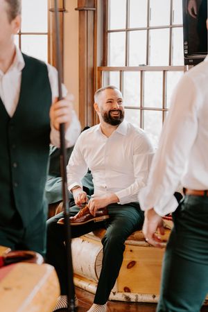 Smiling bearded groom in white shirt and green pants sitting and laughing with groomsmen during wedding preparations.