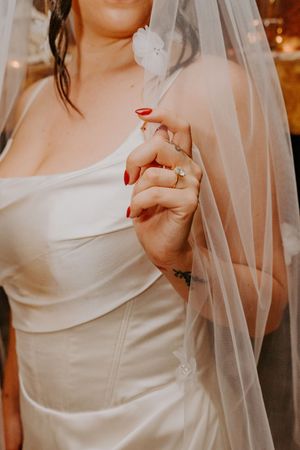 Bride in strapless white satin gown holding sheer veil with red manicured nails and diamond ring.