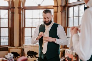 Smiling bearded groom in green vest adjusting his tie while getting ready for his wedding.