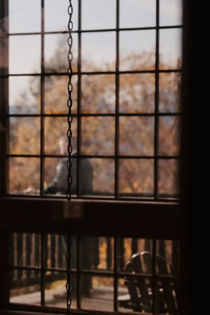 Dark interior view through a grid window with hanging chain, overlooking autumn trees and outdoor porch in warm sunlight.