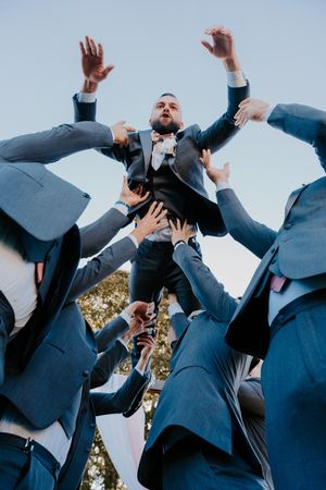 Groomsmen tossing groom into the air in blue suits outdoors during a wedding celebration.