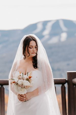 Elegant bride in white dress and pearl veil holds bouquet with snowy mountain backdrop.