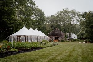 White tent pavilion beside a rustic barn surrounded by lush gardens and trees at an outdoor wedding venue.