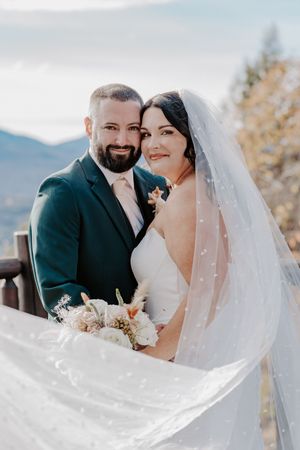 Smiling bride and groom pose with flowing veil and bouquet against mountain scenery.