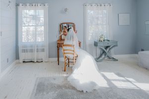 Bride in a white wedding gown and veil sits at a vanity in a sunlit blue room with white floors.