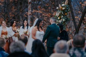 Bride and groom exchange vows at outdoor fall wedding ceremony beneath floral arch in autumn forest.