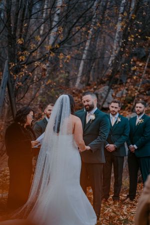 Bride and groom exchange vows at an outdoor fall wedding ceremony in a forest with groomsmen standing nearby.