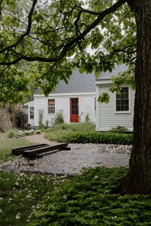 Charming white cottage with red door surrounded by lush green trees and flowering garden landscape.