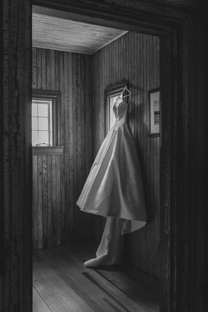 Black and white photo of a wedding dress hanging in a rustic wooden room with hardwood floors and vintage windows.