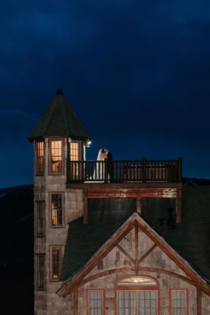 Couple embracing on a lit balcony of a rustic lodge tower at night under a dramatic dark blue sky.