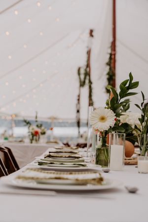 Elegant wedding reception table setting with white gerbera daisies, green charger plates, and glassware under a tent.