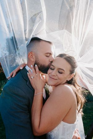 Groom kissing bride on cheek under flowing white veil, bride smiling with eyes closed, wearing wedding dress outdoors.